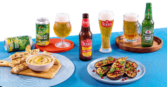 Assortiment van speciaalbieren van Gebrouwen door Vrouwen, Texels en Heineken met twee glazen bier, geserveerd met flatbread en hummus en gegrilde aubergine met granaatappelpitten op een blauw tafelkleed.
