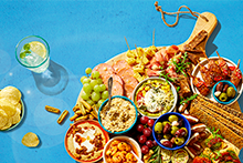 Borrelplank met diverse hapjes zoals druiven, olijven, chips, dips, crackers, vleeswaren, kaas, groenten en een glas frisdrank met citroen en munt.