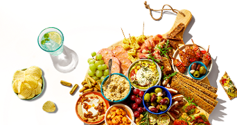 Borrelplank met diverse hapjes zoals druiven, olijven, chips, dips, crackers, vleeswaren, kaas, groenten en een glas frisdrank met citroen en munt.