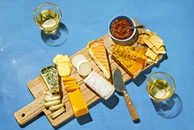 Kaastafel met verschillende soorten kaas, crackers, dip en twee glazen witte wijn, geserveerd op een houten plank.