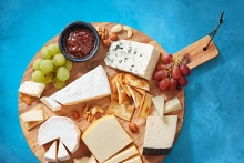 Kaasplank met verschillende soorten kaas, crackers, druiven, noten en een bakje jam op een houten serveerplank.