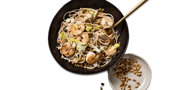 Pasta met garnalen en stukjes avocado, geserveerd in een zwarte kom met een bijgerecht van geroosterde pijnboompitten.