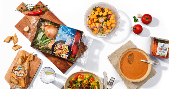 Selectie van Albert Heijn producten en maaltijden, zoals een groentepakket, soep, salade, tomaten, wortelreepjes en stokbrood, op een donkere ondergrond gepresenteerd