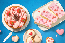 Assortiment van gebak en koekjes met roze en hartvormige decoraties, waaronder een taart met aardbeien, gebakjes met roze glazuur en hartjes, en slagroomsoesjes.