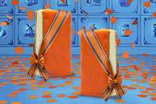 Twee oranjetompoucen met oranje glazuur, gedecoreerd met een lint in de kleuren van de Nederlandse vlag en een oranje strik, op een ondergrond van oranje confetti.