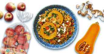 Een blauwe schaal met een couscousgerecht dat pompoen, spinazie, feta en stukjes rode paprika bevat, omringd door AH appels in een zak, losse appels, een halve flespompoen en een assortiment paddenstoelen.