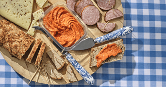 Een houten plank met diverse hapjes, waaronder sneetjes brood, kaas, worst en een schaaltje met rode tapenade, op een blauwe geruite tafelkleed.