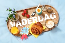 Vaderdag ontbijt met croissant, koffie, zachtgekookt ei, sinaasappelsap en aardbeien op dienblad met bloemen en de tekst "Vaderdag".
