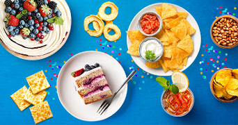 Diverse selectie van feestelijke snacks en desserts, waaronder bessentaarten, tortillachips met dips, gedroogde appelringen, crackers, pinda's en een glas koude drank met citroen en munt, op een blauwe achtergrond.