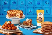 Verschillende typisch Nederlandse lekkernijen, waaronder een stuk appelkruimeltaart met slagroom, een schaal bitterballen, blokjes kaas met prikkers, een stapel stroopwafels, en een fles AH pindakaas op de achtergrond.