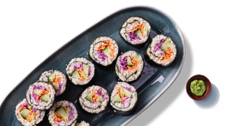 Sushirrolletjes met groenten op een donker ovaal bord, met een bakje wasabi ernaast