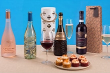 Diverse selectie van flessen wijn en mousserende wijn, met drie gevulde wijnglazen, twee cadeauverpakkingen en een bord met kleine luxe hapjes op een beige tafel.