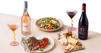 Een sfeervolle gedekte tafel met een fles rosé wijn, een fles rode wijn, glazen wijn, een maaltijd met gegrild vlees en cherrytomaatjes, een verse salade en een borrelplank met diverse kazen, noten, olijven en plakjes appel.