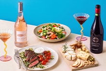 Een sfeervolle gedekte tafel met een fles rosé wijn, een fles rode wijn, glazen wijn, een maaltijd met gegrild vlees en cherrytomaatjes, een verse salade en een borrelplank met diverse kazen, noten, olijven en plakjes appel.
