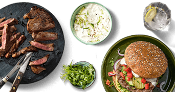 Bord met gegrilde biefstuk, een hamburger met groenten op een volkorenbroodje, kommetje met yoghurtsaus, verse koriander en een glas water met citroen.