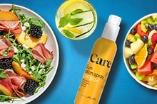 Salade met perzik, prosciutto, bramen en feta op een bord, een glas water met citroen, komkommer en munt, Care zonbeschermingsspray SPF 50+ en een kom met vers fruit zoals meloen, kiwi, bessen en sinaasappel.