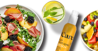 Salade met perzik, prosciutto, bramen en feta op een bord, een glas water met citroen, komkommer en munt, Care zonbeschermingsspray SPF 50+ en een kom met vers fruit zoals meloen, kiwi, bessen en sinaasappel.