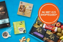 Diverse selectie van ontbijt- en tussendoorproducten, waaronder Perla koffie, een notenreep, biologische citroengemberthee, diverse soorten L'OR koffiecups, Biscoff koekjes, gember, en een blauwe krat gevuld met vers fruit zoals appels, bananen en kiwi’s.