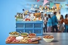 Blauwe Albert Heijn boodschappenkrat gevuld met diverse A-merk producten zoals Tyrrells chips, Bar-le-Duc water en Gran Pavesi crackers, voor een borrelmoment. Voor de krat staan een houten plank met vleeswaren, kaas, crackers, dips en schaaltjes met olijven en nootjes.