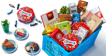 Blauwe Albert Heijn boodschappenkrat gevuld met diverse producten zoals Celebrations chocolade, Merci chocolade, Oatly havermelk, en bananen, met een aparte tafel waarop taart en koffie zichtbaar zijn.