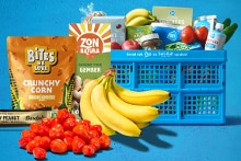 Blauwe Albert Heijn krat gevuld met diverse boodschappen zoals bananen, cherrytomaten, Bites We Love Crunchy Corn, Zonatura groene thee gember, Barbell Salty Peanut reep en andere verse producten.