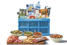 Blauwe Albert Heijn boodschappenkrat gevuld met diverse A-merk producten zoals Tyrrells chips, Bar-le-Duc water en Gran Pavesi crackers, voor een borrelmoment. Voor de krat staan een houten plank met vleeswaren, kaas, crackers, dips en schaaltjes met olijven en nootjes.