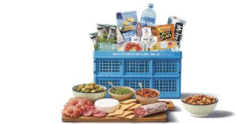 Blauwe Albert Heijn boodschappenkrat gevuld met diverse A-merk producten zoals Tyrrells chips, Bar-le-Duc water en Gran Pavesi crackers, voor een borrelmoment. Voor de krat staan een houten plank met vleeswaren, kaas, crackers, dips en schaaltjes met olijven en nootjes.