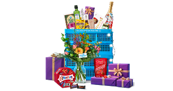 Twee gestapelde blauwe Albert Heijn boodschappenkratten gevuld met diverse boodschappen en versierd met een slinger kerstlichtjes met een kleurrijk boeket bloemen in een vaas ervoor, een doos celebrations, Lindt chocolade en paarse cadeauverpakkingen met goude strikken.