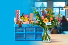 Blauwe Albert Heijn boodschappenkrat gevuld met diverse boodschappen en een kleurrijk boeket bloemen in een vaas ervoor.