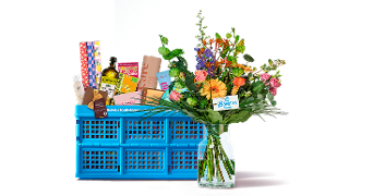 Blauwe Albert Heijn boodschappenkrat gevuld met diverse boodschappen en een kleurrijk boeket bloemen in een vaas ervoor.