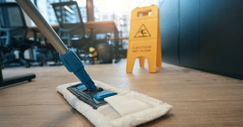 Een werkvloer die wordt gedweild met op de achtergrond bureaus en bureaustoelen en een 'pas op natte vloer' bord