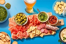 Blauwe Albert Heijn boodschappenkrat gevuld met diverse A-merken en huismerk producten, omgeven door een borrelplank met kaas, vleeswaren, crackers, dips en een schaaltje noten.