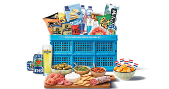 Blauwe Albert Heijn krat gevuld met een variatie aan snacks en boodschappen, waaronder Doritos chips, TUC crackers, Heineken bier, Coca-Cola, Red Bull, kaasblokjes met Nederlandse vlagprikkertjes, toast, verschillende soorten smeersels, noten, en vleeswaren op een houten serveerplank.