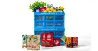 Blauwe Albert Heijn boodschappenkrat gevuld met citroenen, munt, huismerkproducten zoals Perla koffie en Clipperthee. Op de voorgrond pakken Perlakoffie, Clipper thee en Barista Soja.