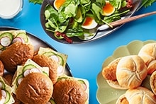 Blauwe Albert Heijn boodschappenkrat gevuld met diverse AH huismerkproducten zoals brood, beleg, kaas, groente en hummus. Voor de krat liggen verschillende soorten meergranenbrood uit de Liefde & Passie lijn, deels gesneden, en een bordje met belegde broodjes met komkommer, tomaat en kruiden.