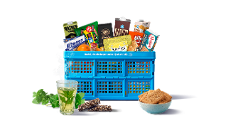 Blauwe Albert Heijn boodschappenkrat gevuld met diverse A-merken en huismerkproducten zoals Nature Valley, B’tween, Kusmi Tea, Barista koffie, ontbijtkoek en vers fruit, met daarnaast een glas muntthee, een kom suiker en mueslirepen op de voorgrond.