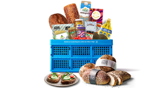 Blauwe Albert Heijn krat gevuld met diverse boodschappen zoals brood, bananen, zuivelproducten, ontbijtgranen en snacks. Voor de krat liggen verse broodjes en een bord met belegde boterhammen.