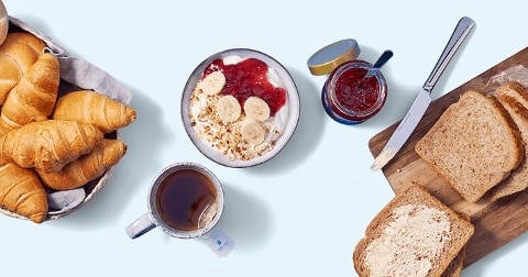 Ontbijt met croissants in een mandje, een kom yoghurt met banaan, granola en aardbeienjam, volkorenbrood met boter en jam op een snijplank, een potje jam, een mes en een kop thee.
