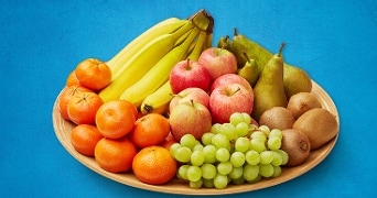 Fruitpalet met bananen, appels, mandarijnen, druiven, kiwi's en peren op een houten schaal voor een blauwe achtergrond.