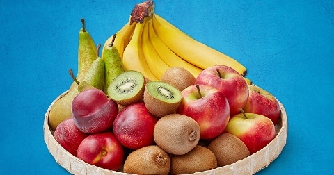 Mand met verschillende soorten fruit, waaronder bananen, kiwi's, peren, pruimen en rode appels, op een blauwe achtergrond.