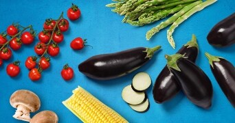 Een assortiment van groenten, waaronder aubergines, kerstomaten, asperges, maïs, en champignons, op een blauwe achtergrond.