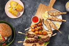 Gegrilde kipsatés, rundvleesbrochettes, maïskolf, en een burger met bijgerechten op een tafel met een drankje en sausjes.