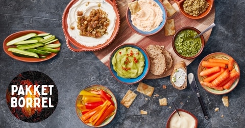 Borrelpakket met diverse dips, crackers, brood, komkommer, wortel en paprika, gepresenteerd op een tafel, met het logo "Pakket Borrel" links in beeld.