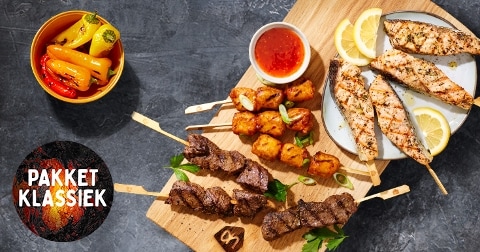 Grillpakket klassiek met verschillende soorten gegrilde spiesen, kipfilet, rundvlees, groenten en dip, gepresenteerd op een houten plank met citroenpartjes en een bakje dipsaus. Op de achtergrond kleurrijke snackpaprika’s en het logo “Pakket Klassiek”.