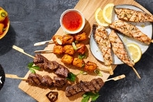 Grillpakket klassiek met verschillende soorten gegrilde spiesen, kipfilet, rundvlees, groenten en dip, gepresenteerd op een houten plank met citroenpartjes en een bakje dipsaus. Op de achtergrond kleurrijke snackpaprika’s en het logo “Pakket Klassiek”.