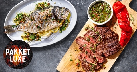 Gegrilde hele vis geserveerd op een schaal met kruiden en citroen, naast gegrilde steak, rode paprika en een bakje groene kruiden saus op een houten plank. Met het label "Pakket Luxe".