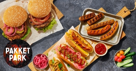 Twee hamburgers met sesambroodjes, twee hotdogs met gegrilde worst, een bakje ketchup, ingelegde uien en verschillende verse groenten zoals cherrytomaatjes en komkommer, gepresenteerd op houten planken. Het woord "Pakket Park" staat links onder in beeld.