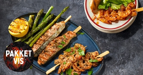 Gegrilde vis- en garnalenspiesen met asperges en citroen, geserveerd op een bord. Naast het bord staat een schaaltje met garnalen en sla. Tekst "pakket vis" zichtbaar op de afbeelding.
