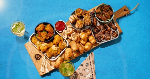 Borrelplank met diverse hapjes zoals mini kroketten, bladerdeeghapjes, gevulde rolletjes, pittige snacks en sausjes, geserveerd op een houten plank met twee drankjes.