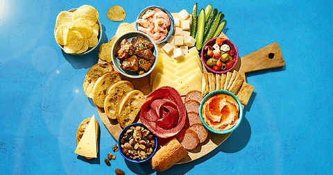 Borrelplank met diverse hapjes op een houten plank, waaronder chips, brood, gemengde noten, verschillende soorten kaas, vleeswaren, salami, komkommer, garnalen, olijven, dips en crackers, op een blauwe achtergrond.